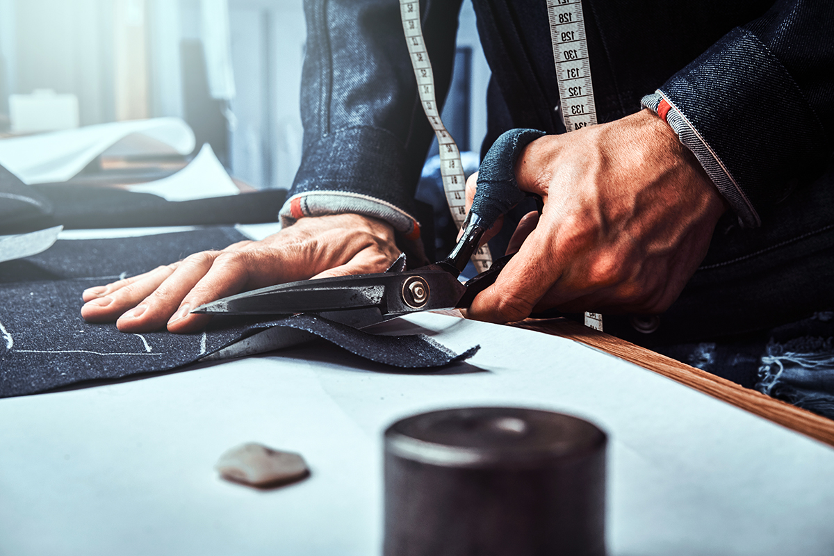 Fashion designer is cutting off fabric with scissors. He is wearing denim. Closeup photo shoot.