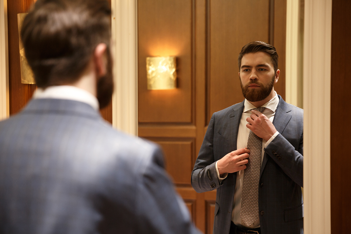 View from back of a young bearded man looking at the mirror and corrects his tie while being in a shop